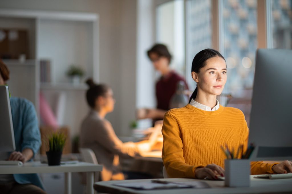 Young pretty businesswoman in yellow pullover sitting by desk and looking at data on computer screen